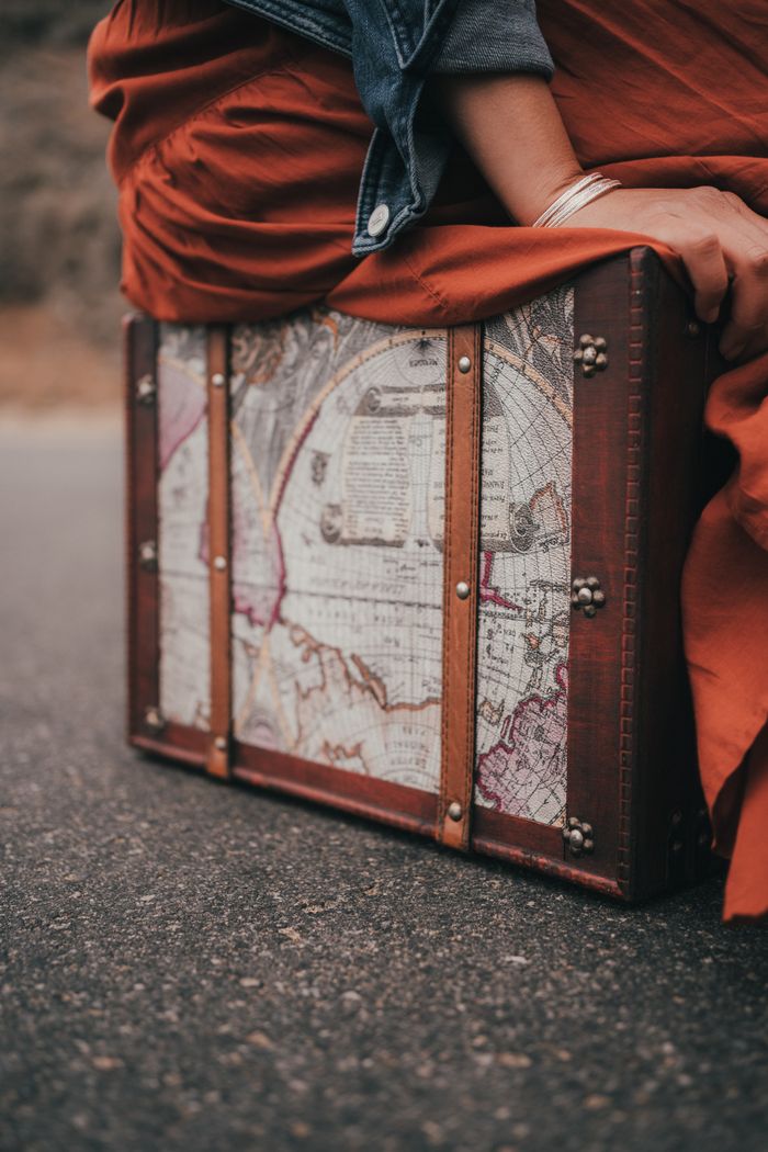gallery-05 A woman sits on a vintage suitcase adorned with an old map design, outdoors.